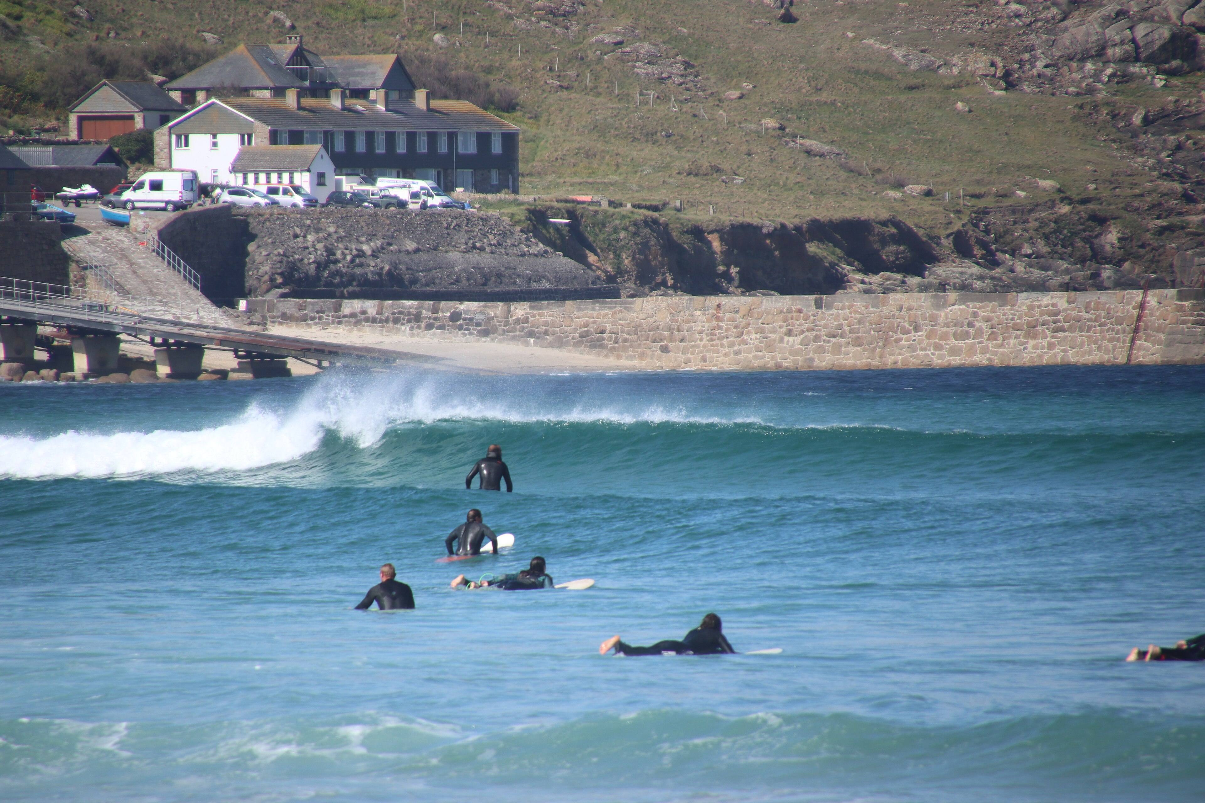 Surfers waiting in the lineup at a Cornish beach — wetsuited figures sitting on boards beyond the break, waiting for the next set.