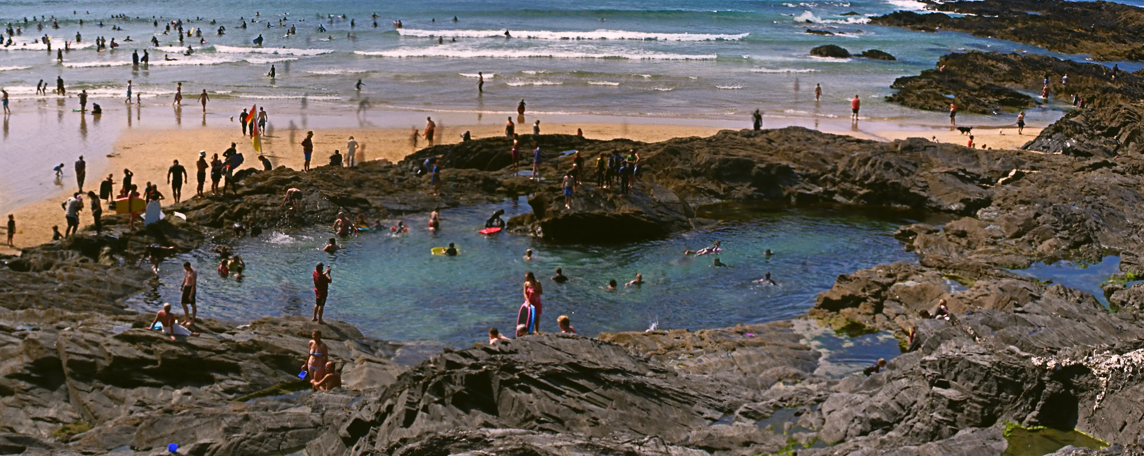 A rock pool at low tide on a Cornish beach — clear water among dark rocks, full of marine life.